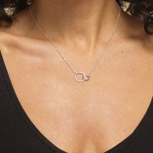 May include: A silver-toned necklace featuring two interlocking circular rings as a central pendant. The delicate chain is secured around the neck, showcasing a polished metallic finish that catches the light against the dark fabric of the top.