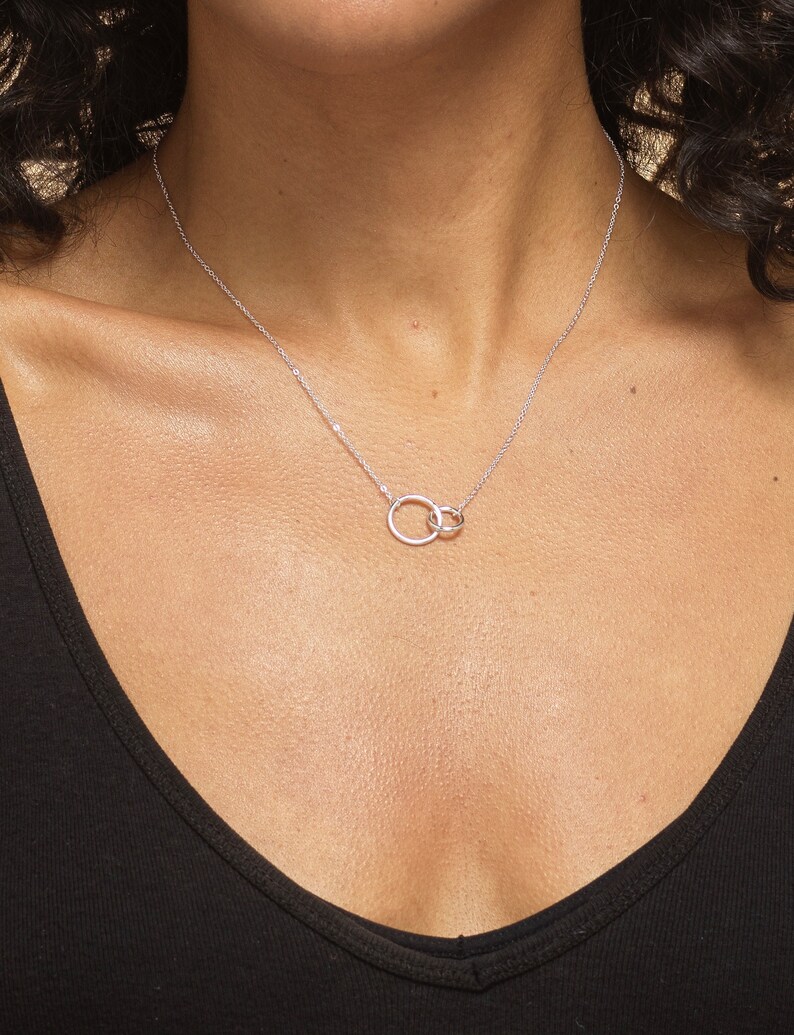 May include: A silver-toned necklace featuring two interlocking circular rings as a central pendant. The delicate chain is secured around the neck, showcasing a polished metallic finish that catches the light against the dark fabric of the top.