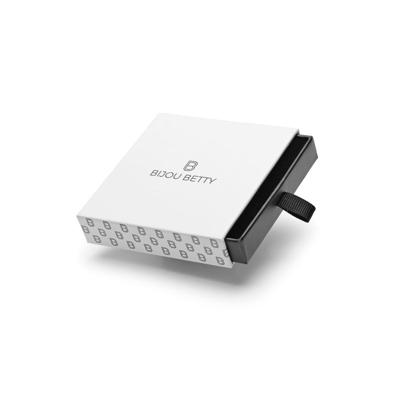 May include: A square white jewelry box with a black sliding drawer and a black fabric pull tab. The top lid features the text BIJOU BETTY with a logo, while the side of the white sleeve is decorated with a repeating pattern of the letter B.
