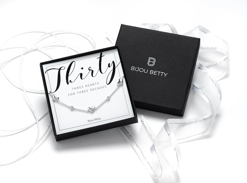 May include: A silver necklace with three heart charms and the text "Thirty Three Hearts for Three Decades" on a white card in a black box. The box has the text "Bijou Betty" on it.