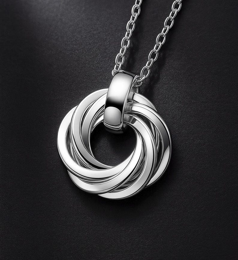 May include: A silver-toned pendant necklace featuring a circular design composed of seven interlocking rings. The pendant is suspended from a matching silver-toned chain, with a polished, reflective metallic finish throughout the entire piece.
