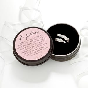 May include: A silver feather ring is displayed in a black-lined, round tin. The tin's lid, with the text "A feather," is open, revealing the ring. White ribbon curls around the tin on a white background. The text on the lid reads "From an angel..."