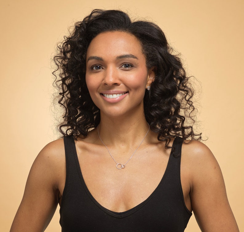 May include: A silver-toned necklace featuring two small, interlocking circular rings as a pendant, suspended on a delicate chain. The necklace is worn against a black tank top, showcasing a simple, polished metal design that rests at the base of the neck.