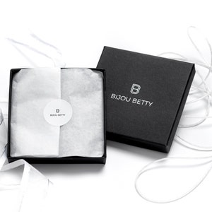 May include: A square black gift box with a matching lid, featuring the white logo and text BIJOU BETTY. Inside, white tissue paper is secured with a circular sticker displaying the same logo and text. White ribbon curls around the box.