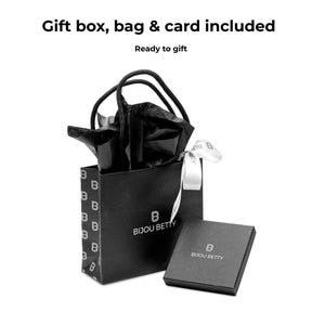 Pode incluir: Uma sacola de presente preta e uma caixa combinando, ambas com o logotipo "Bijou Betty", s&atilde;o apresentadas em um fundo branco. A sacola cont&eacute;m papel de seda preto e uma fita branca. O texto na parte superior diz "Gift box, bag & card included" e "Ready to gift."