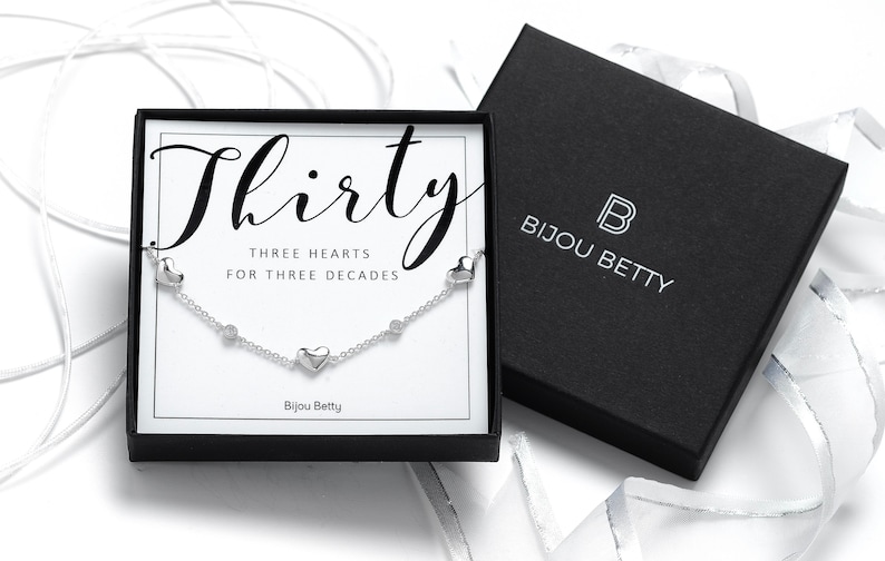 May include: A silver necklace with three heart charms and the text "Thirty Three Hearts for Three Decades" in a black box with the text "Bijou Betty".
