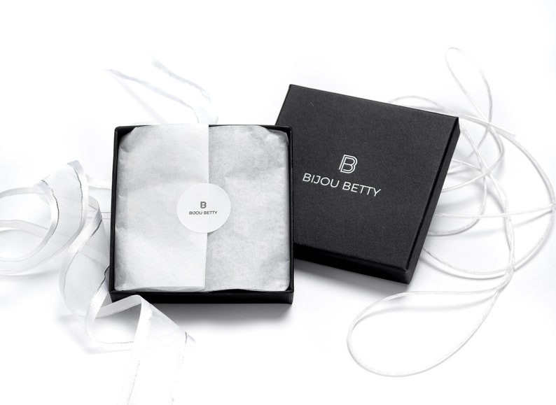 May include: A black gift box with a white logo that says "Bijou Betty" and a white "B" inside a square. The box is open and filled with white tissue paper. A white ribbon is tied around the box.