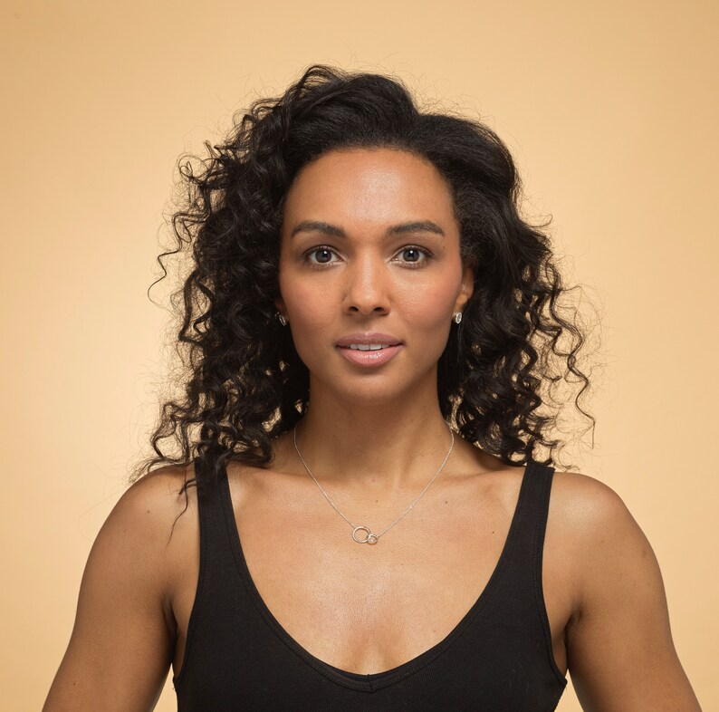 May include: A silver necklace featuring two small, interlocking circular rings as a pendant on a thin chain. The necklace is worn against a black vest top, highlighting the metallic finish and the simple, circular design of the jewellery piece.