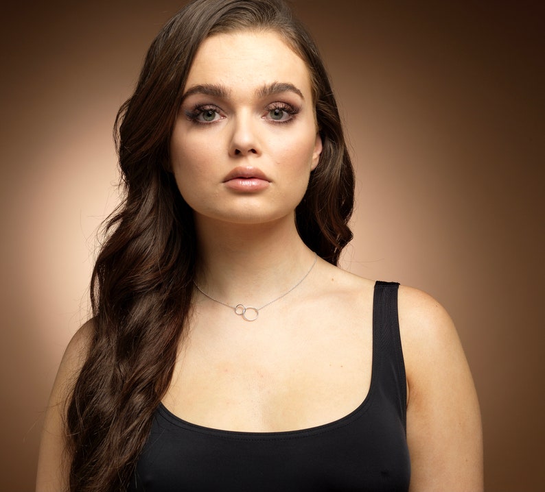 May include: A delicate silver-toned necklace featuring two small, interlocking circular pendants suspended from a thin chain. The necklace rests against the collarbone, showcasing a minimalist design with a polished metallic finish and a simple clasp.