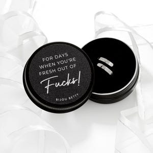 May include: A black round container with the text "FOR DAYS WHEN YOU'RE FRESH OUT OF Fucks!" and the brand name "Bijou Betty". The container is open, revealing a pair of silver earrings. White ribbon is in the background.