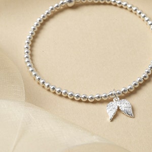 May include: Silver bracelet with small, round beads and a charm shaped like angel wings. The bracelet is made of silver beads. The angel wing charm is detailed and hangs from the centre of the bracelet. The background is a soft, neutral colour.
