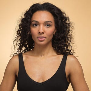 K&ouml;nnte beinhalten: Eine Frau mit dunklem, lockigem Haar und einem schwarzen Tanktop. Sie tr&auml;gt eine zarte goldene Halskette mit einem kleinen Anh&auml;nger. Der Hintergrund ist ein warmer, beigefarbener Farbton.