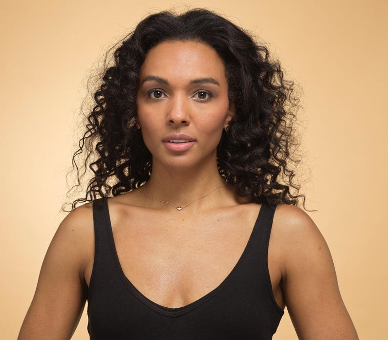 K&ouml;nnte beinhalten: Eine Frau mit dunklem, lockigem Haar und einem schwarzen Tanktop. Sie tr&auml;gt eine zarte goldene Halskette mit einem kleinen Anh&auml;nger. Der Hintergrund ist ein warmer, beigefarbener Farbton.