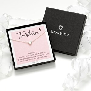 K&ouml;nnte beinhalten: Halskette mit Herzanh&auml;nger in einer schwarzen Schachtel mit dem Markennamen "Bijou Betty". Die Schachtel enth&auml;lt eine rosa Karte mit dem Wort "Thirteen". Wei&szlig;es Band im Hintergrund. Der Anh&auml;nger ist sichtbar.