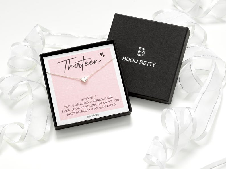 K&ouml;nnte beinhalten: Halskette mit Herzanh&auml;nger in einer schwarzen Schachtel mit dem Markennamen "Bijou Betty". Die Schachtel enth&auml;lt eine rosa Karte mit dem Wort "Thirteen". Wei&szlig;es Band im Hintergrund. Der Anh&auml;nger ist sichtbar.