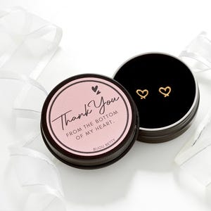 May include: A pair of gold heart-shaped earrings displayed in an open black tin. The tin is next to a closed tin with a pink label that reads "Thank You from the bottom of my heart." White ribbon decorates the background.
