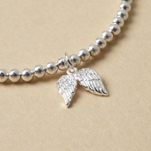 May include: A silver beaded bracelet with a charm of two detailed angel wings. The bracelet features small, round beads and a delicate design. The wings are intricately crafted, adding a touch of elegance to the jewellery.