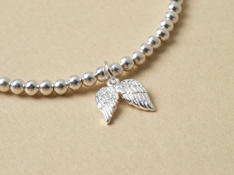 May include: A silver beaded bracelet with a charm of two detailed angel wings. The bracelet features small, round beads and a delicate design. The wings are intricately crafted, adding a touch of elegance to the jewellery.