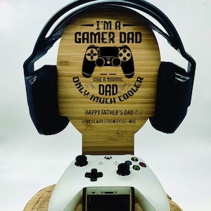 May include: A wooden stand with a gamer dad design. The stand has a cutout for a headset and a space for a video game controller. The stand is engraved with the text "I'm a Gamer Dad, Like a normal dad only much cooler. Happy Father's Day. Love always from Rylee-Mae."