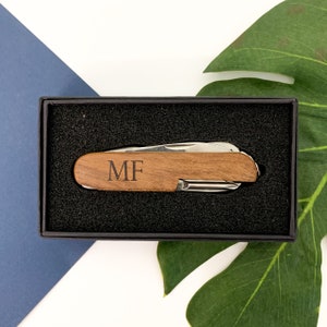 May include: A wooden pocket knife with the initials "MF" engraved on the handle, presented in a black gift box. The knife has a silver blade and tools. The box is set against a backdrop of a blue surface and a green leaf.