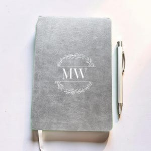 May include: A light grey notebook with a textured cover featuring a white printed design of a circular wreath surrounding the letters MW. A silver metal pen is positioned vertically next to the notebook on the right side.