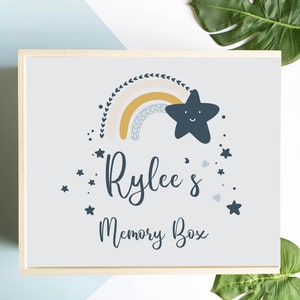May include: A wooden memory box with a light blue background. The box features a rainbow, star, and the text "Rylee's Memory Box" in a whimsical font. The rainbow has yellow, blue, and white colours. Small stars and hearts surround the text.