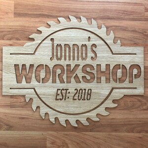May include: A light-colored wooden sign shaped like a circular saw blade with a rectangular center. The sign features the laser-cut text Jonno's WORKSHOP and EST: 2018. The wood grain is visible throughout the surface of the circular saw blade sign.