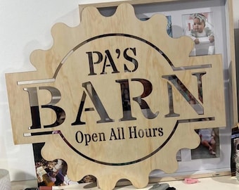May include: A light-coloured wooden sign with a gear-shaped edge featuring the laser-cut text PA'S BARN and Open All Hours in a dark font. The sign has a circular cutout in the centre and is positioned in front of a framed photo collage.