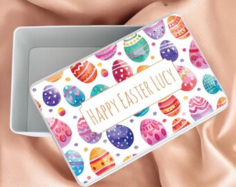 May include: White rectangular tin box with a lid. The lid has a colorful pattern of watercolor Easter eggs with the text "Happy Easter Lucy" in gold lettering.