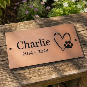 May include: A rectangular copper-coloured metal plaque featuring the engraved name Charlie, the years 2014-2024, and a graphic of a heart with a paw print inside. The plaque has two small circular holes on the left and right sides for mounting.