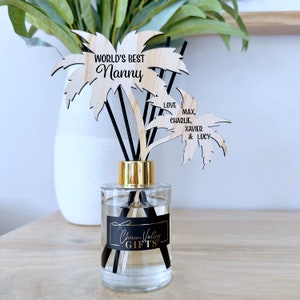 May include: A clear glass diffuser bottle with a gold lid and black diffuser sticks. The bottle has a black label that says "Chain Valley Gifts". The diffuser is topped with two wooden palm tree cutouts. One cutout says "World's Best Nanny" and the other says "Love Max, Charlie, Xavier & Lucy".