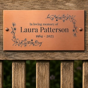 May include: A rectangular copper-toned memorial plaque featuring black engraved text that reads In loving memory of Laura Patterson 1964 - 2023. The plaque is decorated with black line art of floral vines and secured with two metal screws.