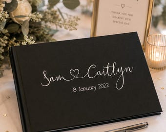 May include: A black rectangular guest book with white text on the cover that reads Sam and Caitlyn with a heart symbol connecting the names and the date 8 January 2022 below. A silver pen lies on the surface in front of the closed book.