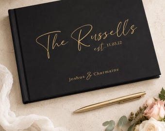 May include: A black rectangular guest book with gold foil lettering that reads The Russells est. 11.03.22 and Joshua & Charmaine. A gold pen is placed diagonally across the bottom right corner of the book cover.