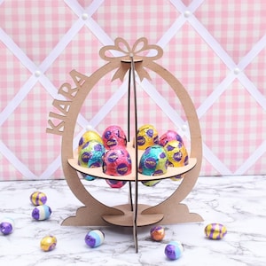 May include: Wooden Easter egg stand with the name "KIARA" written on it. The stand has a bow at the top and holds several colourful chocolate eggs.