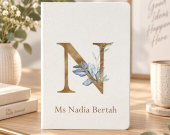 May include: A white hardcover journal featuring a large gold letter N on the front cover, decorated with blue and green watercolor leaves. The name Ms Nadia Bertah is printed in gold text below the letter N on the front cover.