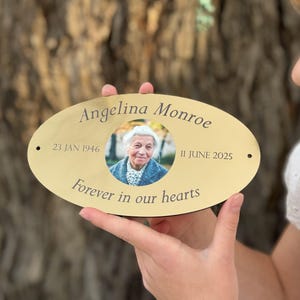 May include: An oval gold-coloured metal plaque featuring a central circular photograph of an elderly person. The text reads Angelina Monroe, 23 JAN 1946, 11 JUNE 2025, and Forever in our hearts. The plaque has two small holes on the left and right sides.