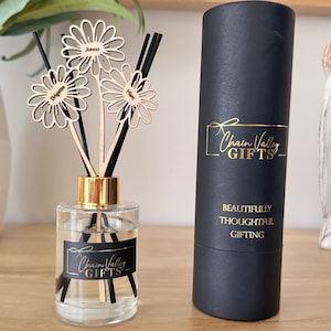 May include: A black diffuser bottle with a gold lid and black diffuser sticks. The bottle has a label that says "Chain Valley Gifts". The bottle is sitting on a wooden surface. There are three wooden flower-shaped tags with names on them. The tags are attached to the diffuser sticks. The diffuser bottle is in front of a black tube with a gold label that says "Chain Valley Gifts" and "Beautifully Thoughtful Gifting".