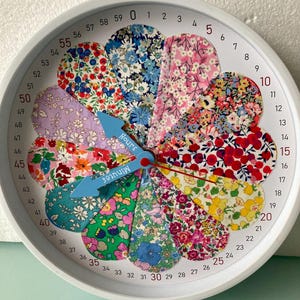 Peut inclure: Une horloge murale ronde avec un cadre blanc et un cadran composé de douze pétales en tissu floral distincts aux couleurs variées. Le bord extérieur affiche les chiffres de 0 à 59. Deux aiguilles bleu clair portent les mentions Hours et Minutes.