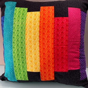 May include: Square decorative pillow with a patchwork design. The pillow features vertical stripes in a rainbow of colors, including blue, green, yellow, orange, red, pink, and purple. The background and borders are black.