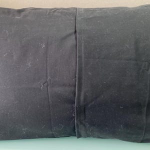 May include: A rectangular black pillow with a seam down the middle. The pillow is made of a dark fabric and appears to be filled with a soft material. The pillow is designed for comfort and support.