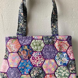 May include: A tote bag constructed from a patchwork of hexagonal fabric pieces featuring various floral and botanical patterns in shades of pink, blue, purple, green, and black. The bag includes two matching fabric handles and purple piping.