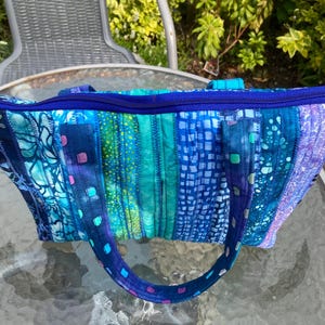 以下が含まれることがあります： 青、ターコイズ、紫色の生地を組み合わせた、キルト加工のトートバッグ。バッグはジッパー開閉式で、お揃いのハンドルが付いています。様々な模様と質感が組み込まれており、ユニークでカラフルなアクセサリーです。