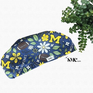 May include: A navy blue scrub cap with a floral and football design. The cap features yellow flowers, white daisies, green leaves, and brown footballs. The word "Wolverines" is printed on the cap, along with the letter "M".