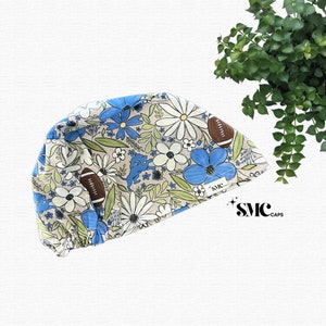 May include: A beige surgical scrub cap with a floral pattern of blue and white flowers, green leaves, and brown footballs. The cap has a curved design and features the text "SMC CAPS" in black.