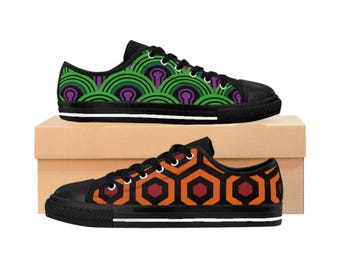 May include: Two pairs of black canvas sneakers. One pair features a green and purple repeating pattern, while the other has an orange and red geometric design. Both pairs have black soles and white midsoles, sitting on a brown cardboard box.