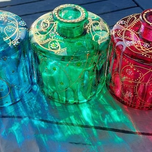May include: Three decorative glass bottles in teal, green, and red. Each bottle has a round body and a narrow neck, with gold and white painted floral designs. The bottles are sitting on a light-coloured surface, with shadows cast by the sunlight.