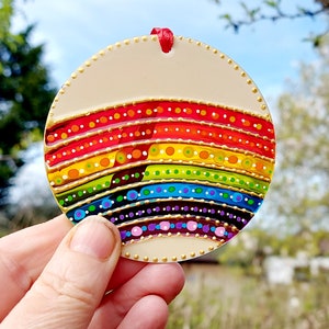 May include: A circular, cream-coloured ornament with a rainbow design. The rainbow features bands of red, orange, yellow, green, blue, and purple, each with white dots and gold accents. A red ribbon is attached for hanging.