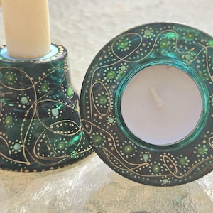 May include: A teal candle holder and a tealight holder with a matching design. Both are decorated with gold and white swirling patterns and green accents. The candle holder has a white candle inserted.