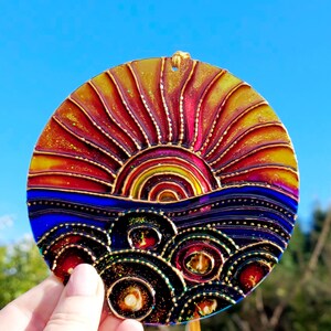 May include: A circular suncatcher featuring a sunset design. The sun is rendered in red, yellow, and orange, above a blue sea. The suncatcher has a gold outline and a small hole at the top for hanging. The diameter is approximately 10 cm.